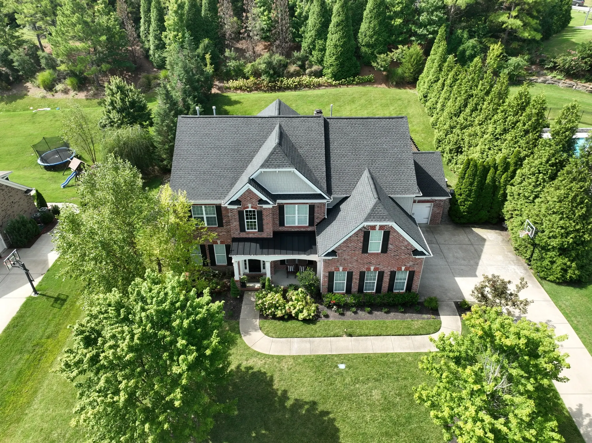 Aerial view of a two-story brick house with a dark roof, surrounded by greenery and a driveway, located in a suburban neighborhood. A trampoline is visible in the backyard.