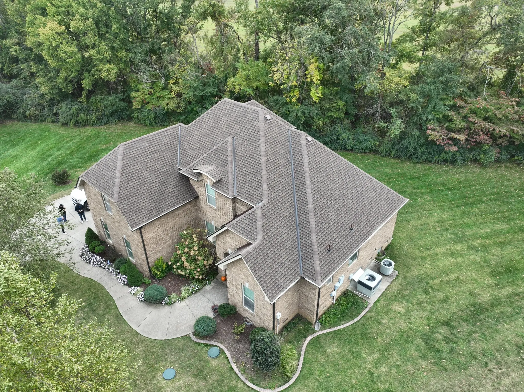 Aerial view of a brick house with a brown roof, surrounded by a green lawn and trees. A curved path leads to the entrance.