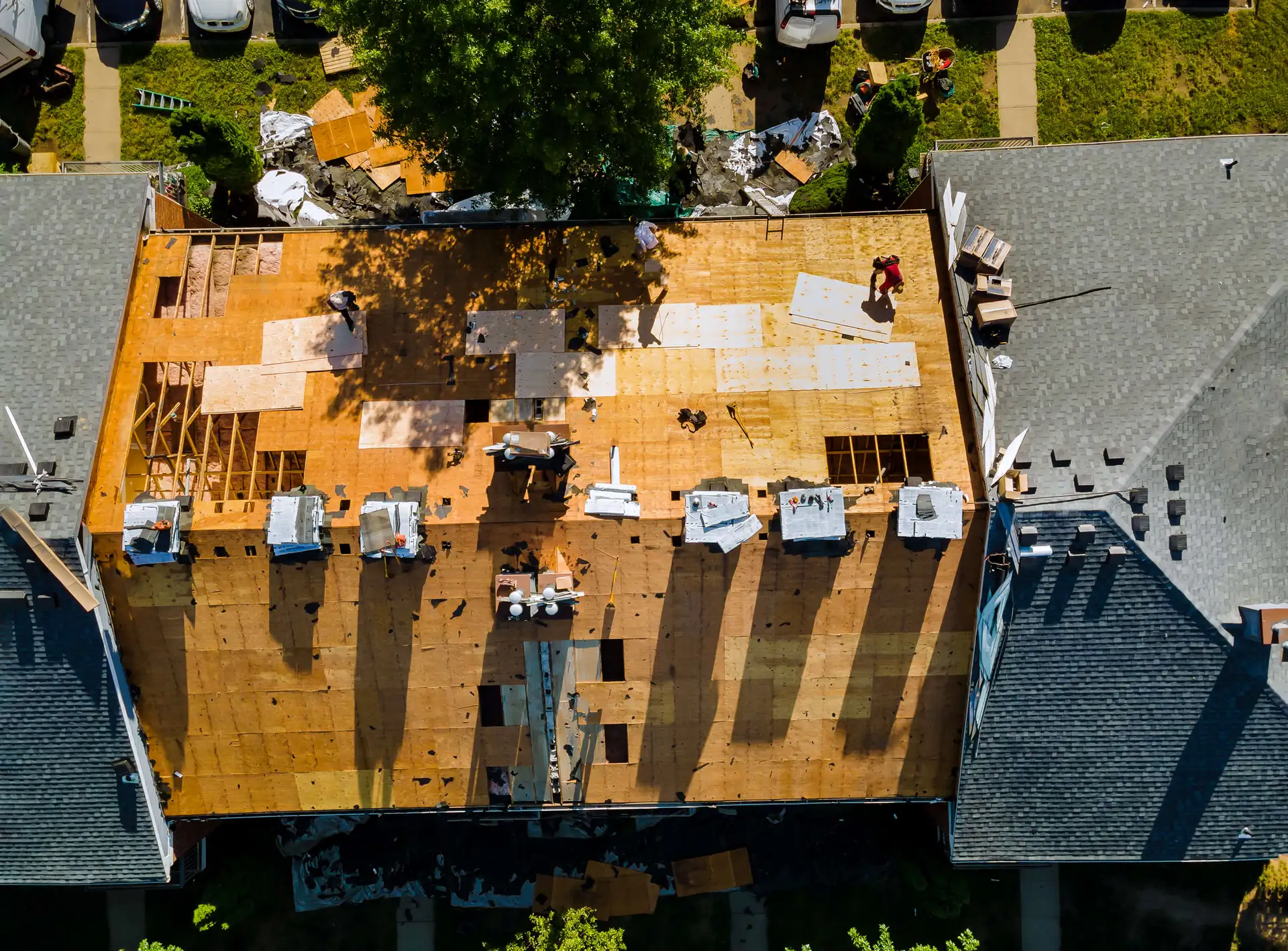 Aerial view of workers repairing a large roof, with tools and materials scattered, and part of the roof covered in new plywood.