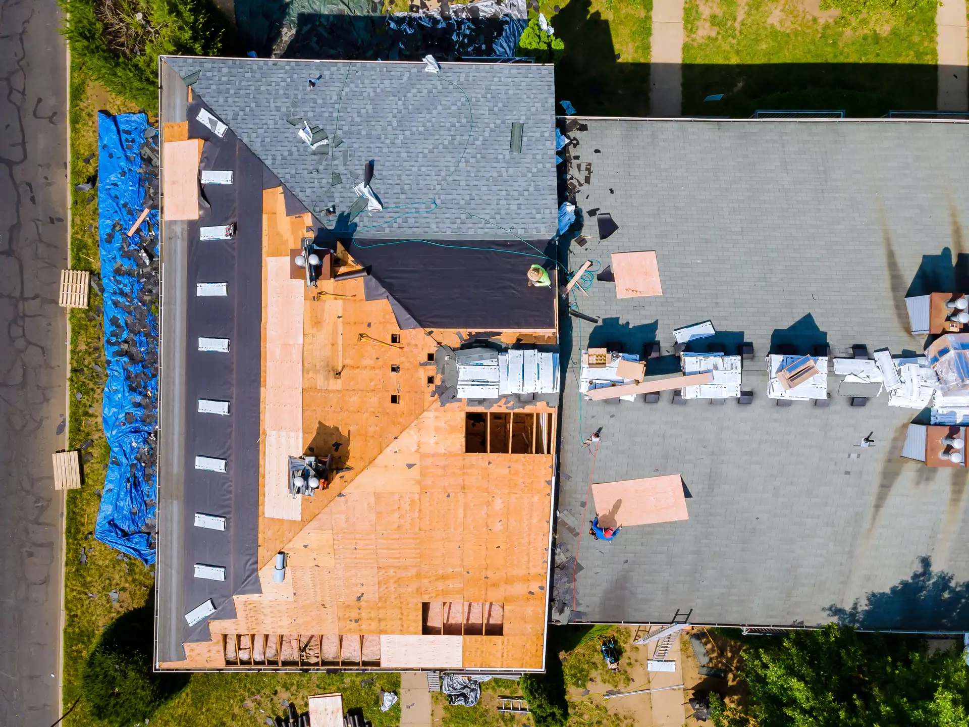 Aerial view of two adjacent rooftops; one is partially stripped, exposing wooden decking, while another is fully shingled. Workers and equipment are present on both roofs.