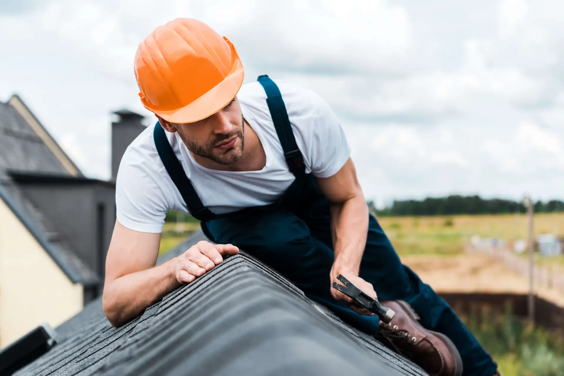 A construction worker wearing an orange hard hat and overalls kneels on a roof while holding a hammer.