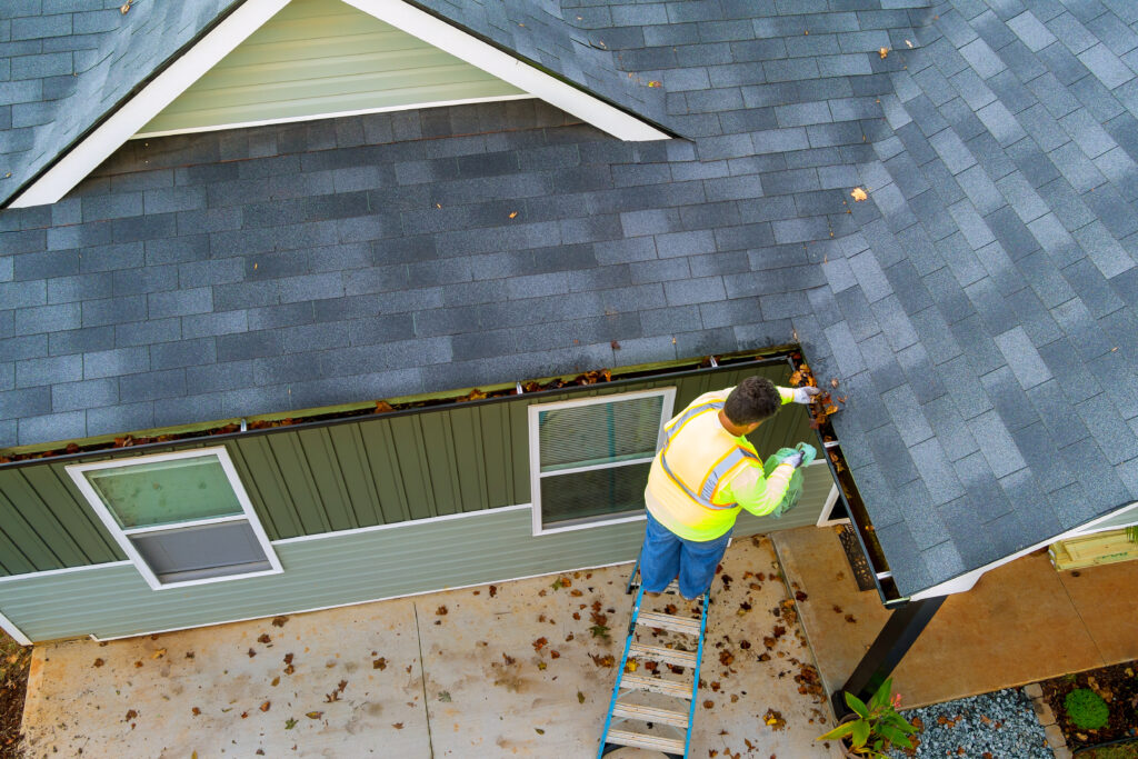 A man in a high-visibility vest stands on a ladder, cleaning leaves from the gutter of a house with grey shingles.