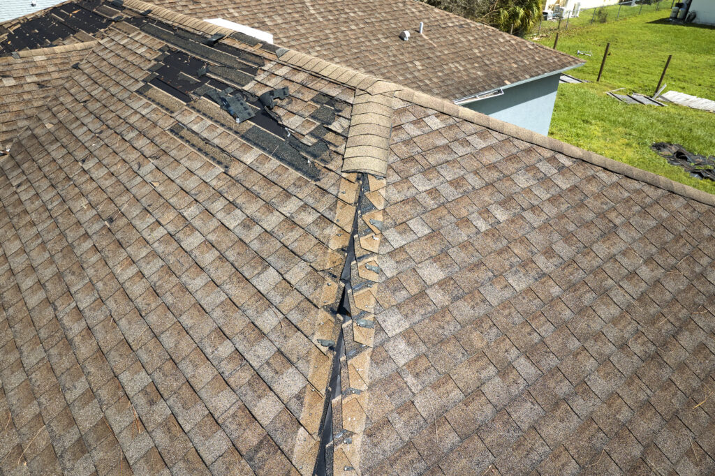 Roof with damaged shingles along the ridge, exposing underlying layers. Some shingles are missing, revealing black material underneath. Surrounding rooftops and grassy area visible.