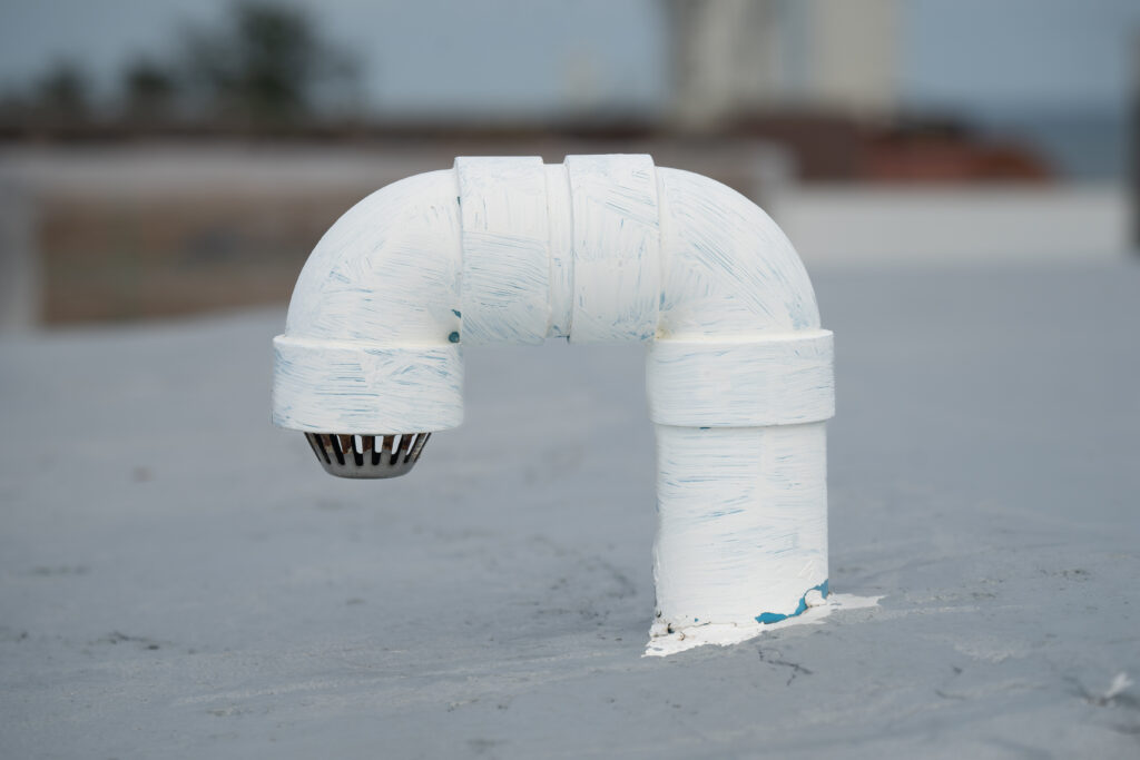 A white-painted rooftop pipe vent with a drain cover is on a flat gray surface.