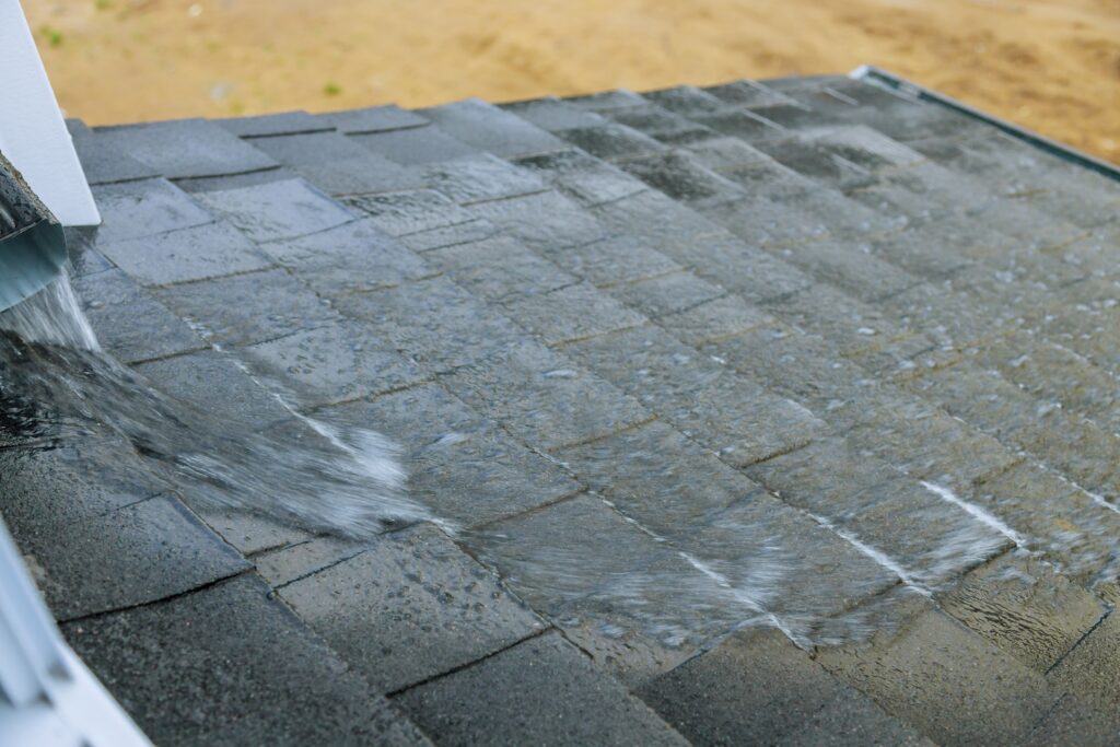 Rainwater flows down gray roof shingles near a metal gutter under an overcast sky.