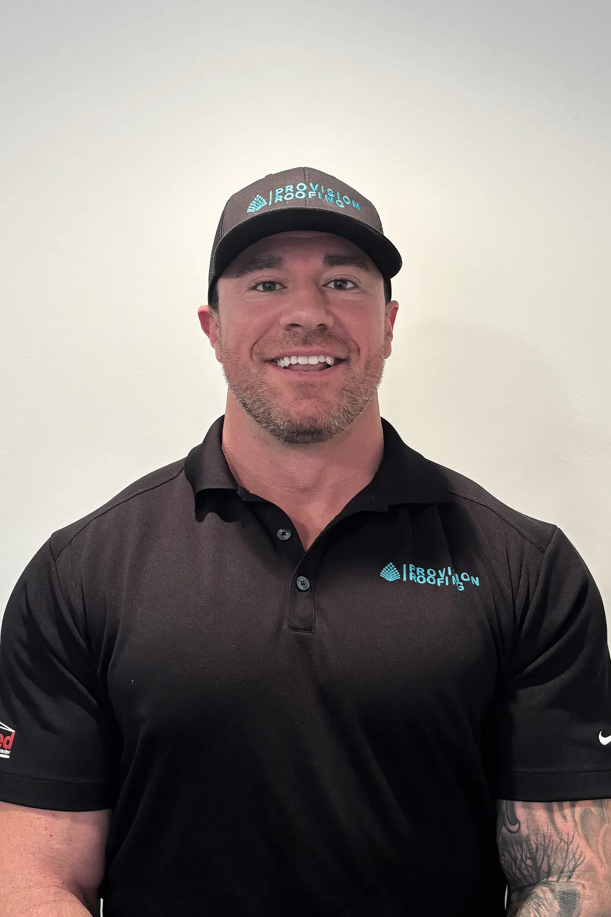 A man wearing a black polo shirt and cap with a logo, standing against a plain white background.